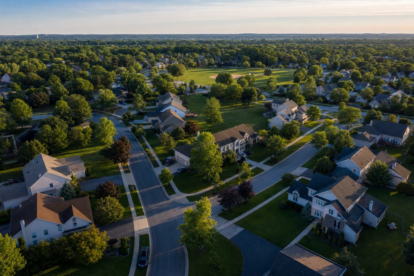 An aerial view of a suburban neighbourhood with varied rooflines and tree canopy at golden hour